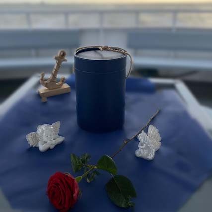 Ein blaue Seeurne, eine rote Rose und Engelfiguren auf einem Tisch mit Seeblick.