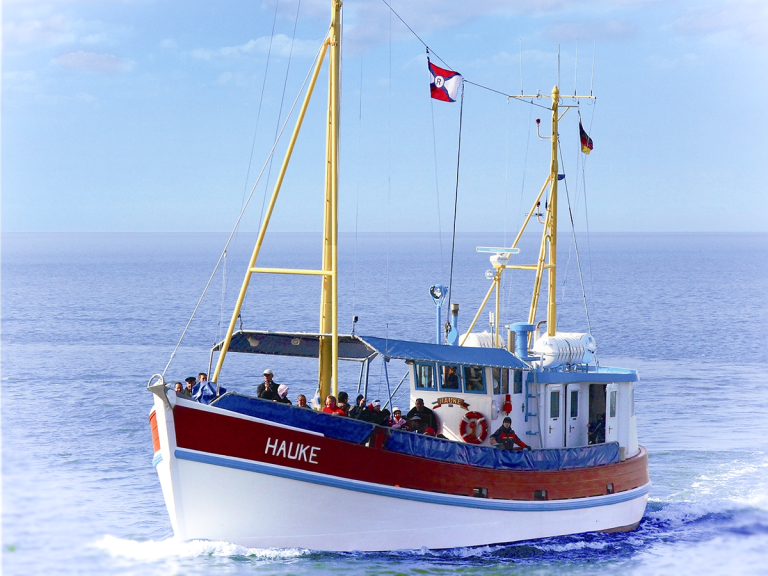 MS Hauke mit roten und blauen Details, fährt über ruhige See mit Flaggen.