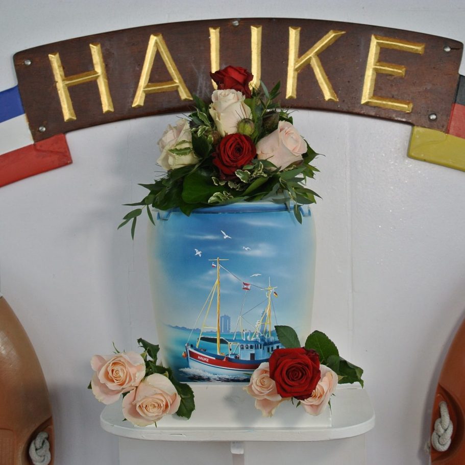 Seeurne mit maritimer Malerei und roten sowie rosa Rosen auf einem Regal.